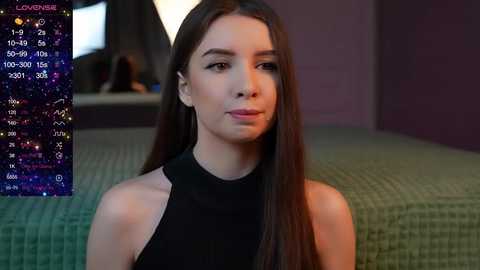 Media: A video of a young woman with long, straight brown hair, wearing a black sleeveless top, sitting on a green textured couch in a dimly lit room. A digital astrological chart overlays her face.