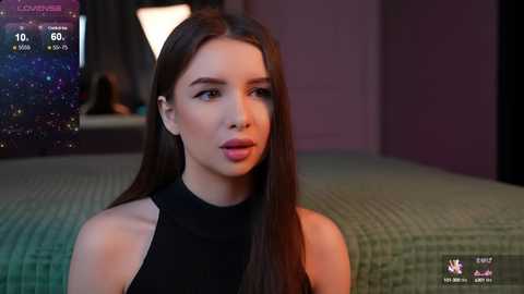 Media: A video of a young woman with long brown hair, fair skin, and pink lips, wearing a black sleeveless top, in a dimly lit bedroom with a green quilted bedspread.