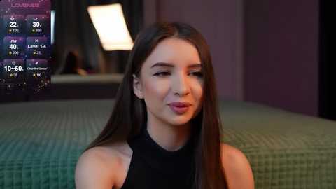 Media: Video of a young woman with long brown hair, wearing a black sleeveless top, sitting on a green quilted bed. Background shows a dimly lit room with a digital clock displaying 22:30.