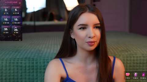 Media: Video of a young woman with long brown hair, fair skin, wearing a blue spaghetti-strap top, sitting indoors on a green-patterned couch.