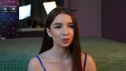 Media: Video of a young woman with long brown hair, wearing a blue spaghetti-strap top, standing in a dimly lit room with a green-patterned bedspread in the background.