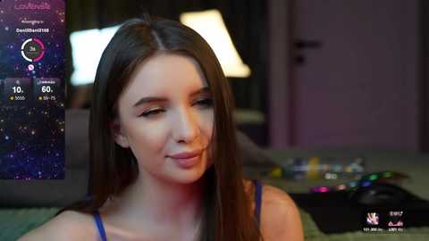 Media: Video of a young woman with long, dark hair, wearing a blue top, smiling slightly, in a dimly lit room with a digital clock and colorful objects in the background.