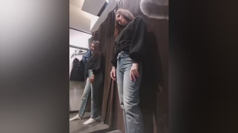 Video of a woman with long brown hair, wearing a black top and high-waisted jeans, leaning against a fitting room wall, with two other women in the background.