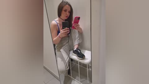 Video of a young woman with straight, shoulder-length brown hair, wearing a black choker, striped pajamas, and black sneakers, taking a selfie in a small, plain bathroom mirror.