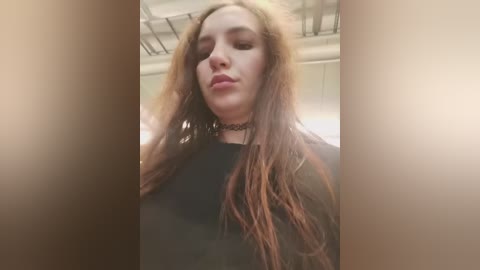 Video of a young woman with long, tousled brown hair, wearing a black choker necklace, black top, and a contemplative expression, captured in a dimly lit room with white walls and a metal ceiling.