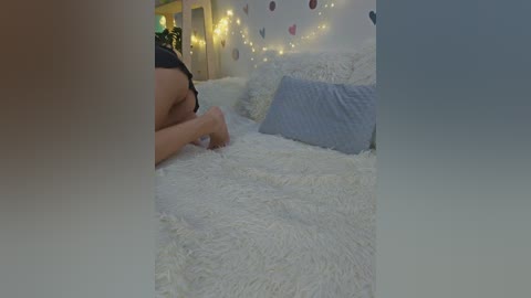Video of a man with a light skin tone, lying face down on a white, fluffy bed, wearing a black shirt. The room has a cozy, dimly lit ambiance with fairy lights and a decorative wall.