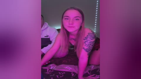 Media: Video of a smiling, topless woman with long blonde hair and tattoos, on all fours on a bed with patterned sheets, in a dimly lit room with purple lighting.