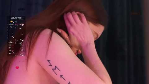 A video of a nude woman with fair skin, long brown hair, and a tattoo of musical notes on her left arm, covering her face with her right hand, dimly lit, blurry background.