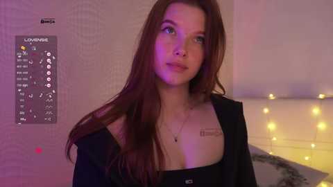Video of a young woman with long, straight auburn hair, light skin, and green eyes, wearing a black top with a low neckline, standing indoors with a calendar and string lights in the background.