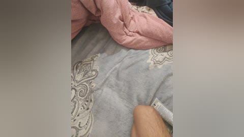 Video of a person lying on a bed with light gray bedding featuring intricate paisley patterns. The person's arm, partially visible, is resting on the bed. The background shows a pink pillow and a dark-colored blanket.