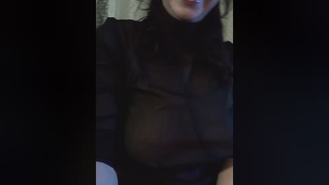 Video of a woman in a sheer black blouse, revealing her large breasts beneath. She has light skin and dark hair. The background is dimly lit, with a blurred, textured wall.