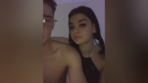 Video of a young woman with long black hair, wearing a black top, standing close to a shirtless man with glasses in a dimly lit room.