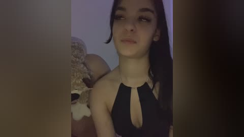 Video of a young woman with fair skin, dark hair, and wearing a black halter top, standing next to a stuffed bear in a dimly lit room.