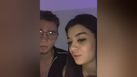 Video of two young people, a man with glasses and a woman with long dark hair, smiling at the camera. The background is dimly lit and out of focus.