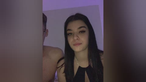 Video of a young woman with long, straight black hair, light skin, and a slender build, wearing a black halter top. She smiles slightly, standing in front of a purple-lit, white-walled room with a person's arm partially visible on the left.