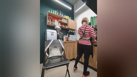 Video of a small, modern barbershop with a blonde woman in a striped shirt and black pants working at a wooden counter. Shelves hold bottles, a black chair, and a white sanitizer dispenser are visible.