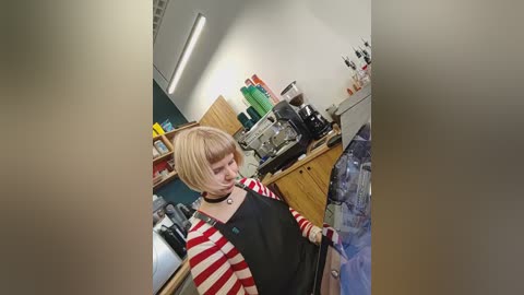 Video of a young person with short blonde hair, wearing a black apron over a red-striped sweater, standing in a cluttered, brightly lit kitchen with wooden cabinets and various appliances.