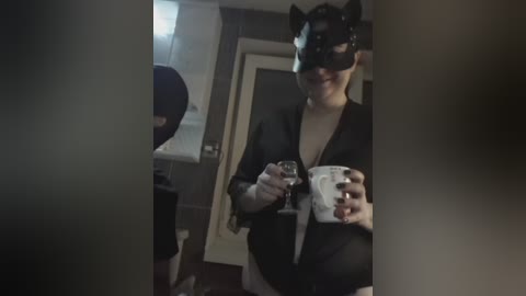 Video of a person in a black cat mask, black suit, and white shirt, holding a white mug, in a dimly lit, modern bathroom with a white door and towel rack.
