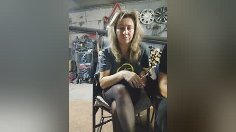 Media: Video of a middle-aged woman with shoulder-length blonde hair, sitting in a workshop with a cluttered background featuring tools, machinery, and a bicycle.