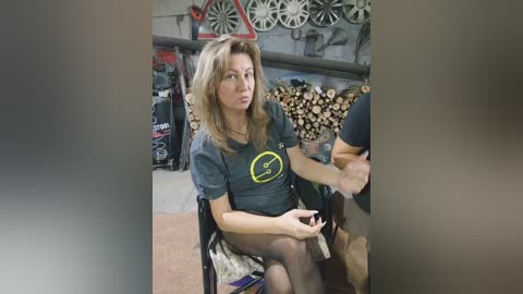 Media: Video of a blonde woman with a medium build, wearing a dark T-shirt with a yellow lightning bolt and black tights, seated on a chair in a cluttered garage.