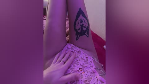 Video of a woman with light skin and a large tattoo of a wolf's head on her thigh, lying on a bed with a pink lace blanket, wearing a silver chain necklace.