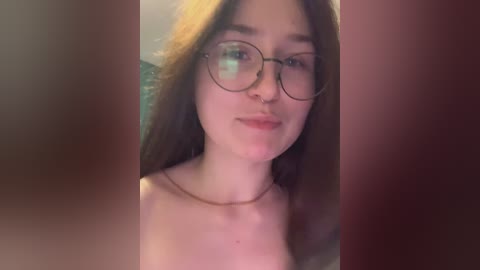 A video of a young woman with fair skin, long brown hair, and large round glasses, smiling slightly. She's wearing a thin necklace. The background is blurred, with a reddish tint.