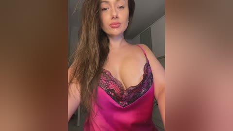 Media: Video of a light-skinned woman with long brown hair, wearing a pink satin camisole with black lace trim, revealing cleavage, indoors with blurred background.
