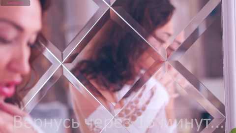 Media: Video of a woman with long, wavy brown hair, wearing a white dress with a beaded necklace, seen through a diamond-patterned glass, with blurred background.
