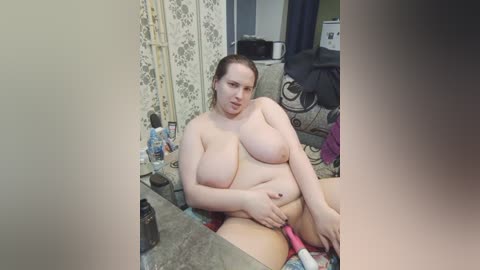 Video of a plus-sized woman with large breasts, sitting naked at a vanity, holding a pink vibrator, surrounded by toiletries and a floral-patterned screen.