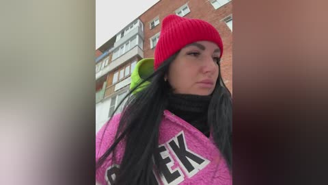 Video of a woman with long black hair, wearing a bright red knit beanie, a pink sweatshirt with black letters, and a black scarf, standing against a brick building background.