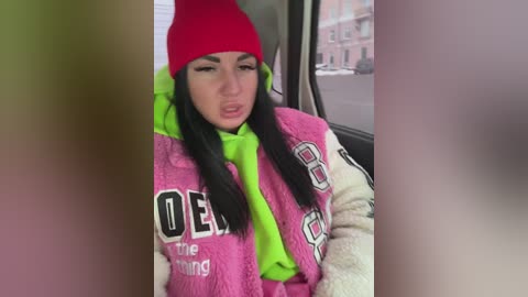 Video of a young woman with long black hair, wearing a bright pink jacket, neon green scarf, and a red beanie, sitting in a car with a blurred cityscape background.