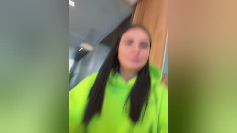 Video of a woman with long black hair and a bright green hoodie, blurred background of an indoor setting with wooden and metal elements.