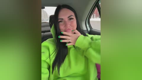 Video of a young woman with long black hair, wearing a bright green hoodie, sitting in a car. She has a playful expression, holding her hand to her face.