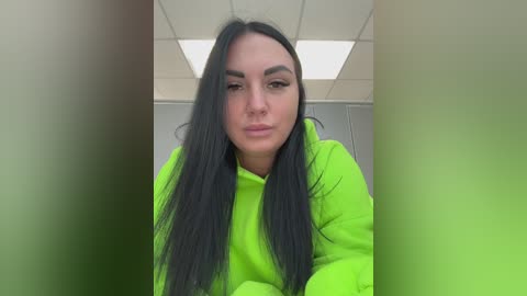 Video of a woman with long black hair, wearing a neon green hoodie, looking directly at the camera. The background shows a white ceiling with fluorescent lights.