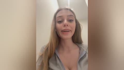 Media: A video of a young Caucasian woman with long, blonde hair, fair skin, and a slim build, wearing a grey shirt with the collar open, smiling in a bathroom with beige walls and a white ceiling.