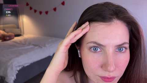 Video of a woman with fair skin and blue eyes, touching her hair, in a bedroom with a bed, heart garland, and a lamp.