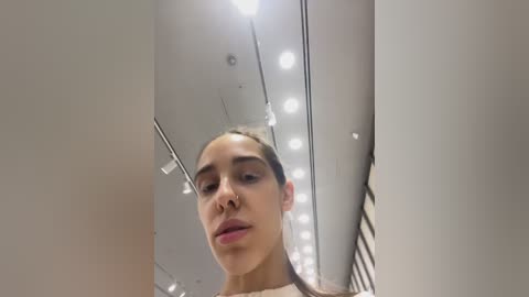 Media: A video of a young woman with light brown skin and dark hair, taken from a low angle, looking up at a ceiling filled with bright lights in a modern indoor space.