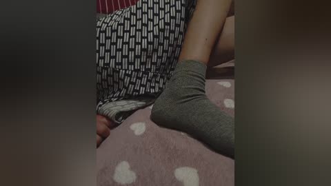 Media: Video of a person in a striped pajama top, lying on a pink, heart-patterned blanket. Their legs, clad in grey socks, are crossed. The background is dimly lit.