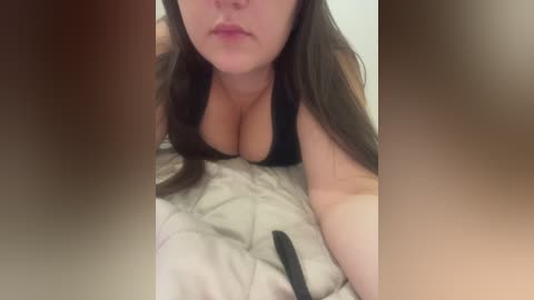 A close-up video of a woman with long brown hair and large breasts in a black bra, lying on a bed with beige sheets. Her face is partially obscured by out-of-focus hands.