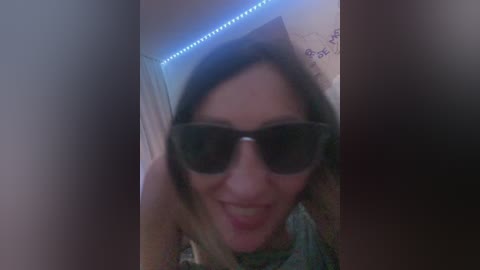 A blurry video of a woman with short brown hair, wearing sunglasses and a floral shirt, standing in a dimly lit room with blue LED lights.