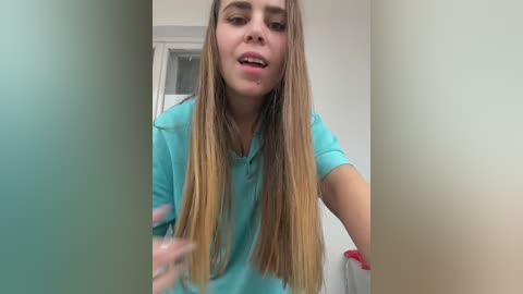 Media: A video of a young woman with long, blonde hair, wearing a teal T-shirt, standing in front of a blurred, beige wall. Her mouth is open, suggesting surprise or excitement.