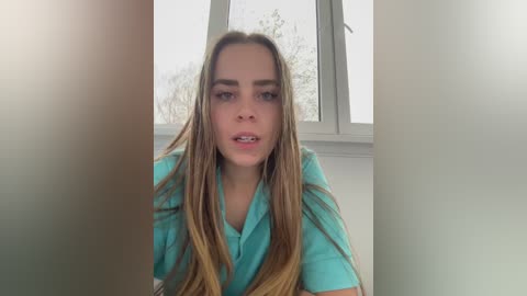 Media: Video of a young Caucasian woman with long, straight, light brown hair and fair skin, wearing a light blue polo shirt, seated indoors near a window with a blurred view of winter trees outside.
