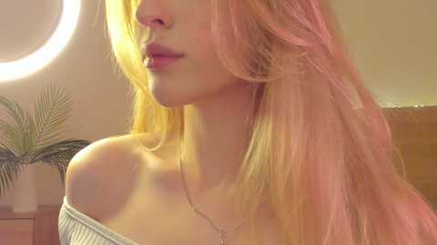 Media: Video of a young woman with long, pastel pink hair, fair skin, wearing a white off-shoulder top and a delicate necklace, set against a soft, warm-toned background with a round light fixture.
