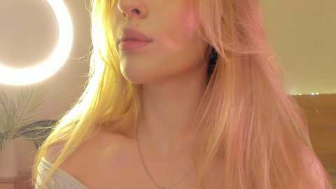Media: A video of a young woman with long, straight blonde hair, fair skin, and full lips. She wears a delicate necklace, and a soft light illuminates her face. The background shows a blurred, warm-toned setting.