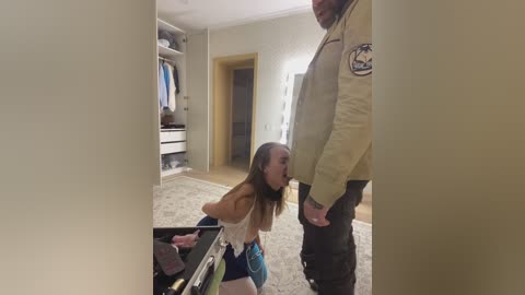 Media: Video of a woman kneeling on a carpeted floor in a beige room, with a man in a beige jacket standing above her.