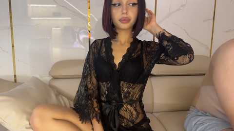 Media: Video of a young woman with short dark hair, wearing a sheer black lace robe, sitting on a beige couch in a modern living room with white marble walls and gold accents.