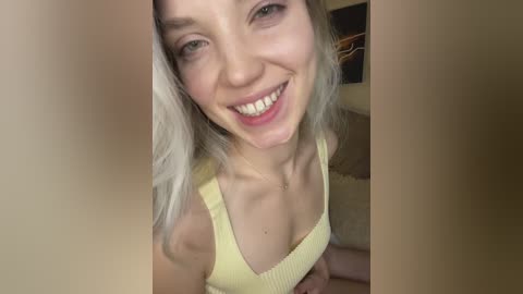 Media: Video of a young Caucasian woman with long, platinum blonde hair and fair skin, smiling broadly. She wears a yellow tank top revealing a hint of cleavage. Background features a blurred abstract painting.