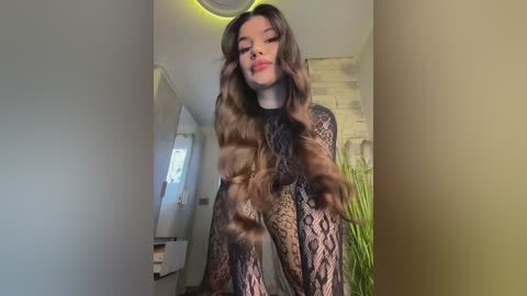 Media: Video of a young woman with long, wavy brown hair, wearing a black, lace bodysuit, standing indoors with a brick wall and potted plants in the background.