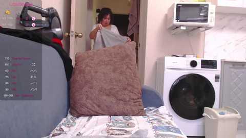 Video of a woman in a small laundry room, holding a brown pillow. Background includes a washer, dryer, microwave, and newspaper-covered table.