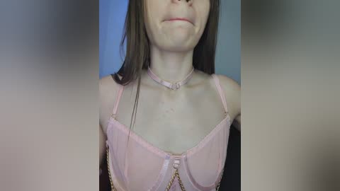 Media: A close-up video of a young woman with pale skin and straight brown hair, wearing a pink bra with thin straps and a delicate gold chain choker. The background is blurred, focusing on her upper body and face.